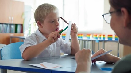 child with Down syndrome at school, pupil boy with disabilities studying mathematics with her female teacher sitting at a desk in classroom - Powered by Adobe