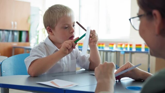 child with Down syndrome at school, pupil boy with disabilities studying mathematics with her female teacher sitting at a desk in classroom