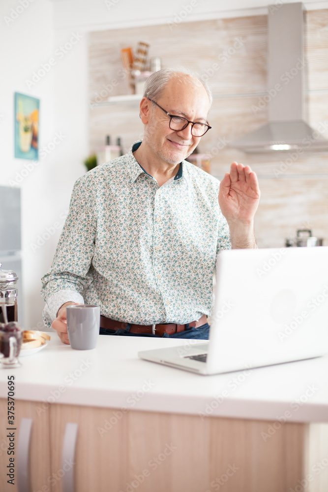 Poster old man saying hello during video call with family using notebook in kitchen while holding a cup of 