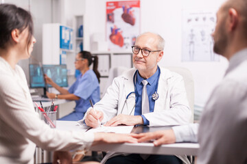 Obraz premium Senior doctor writing notes on clipboard during examination of young couple with fertility illness in hospital office and nurse holding patient x-ray.