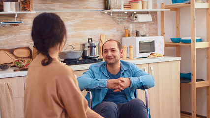 Portrait of happy disabled man in wheelchair looking at wife in kitchen. Guy with paralysis handicap disability handicapped difficulties getting help for mobility from love and relationship