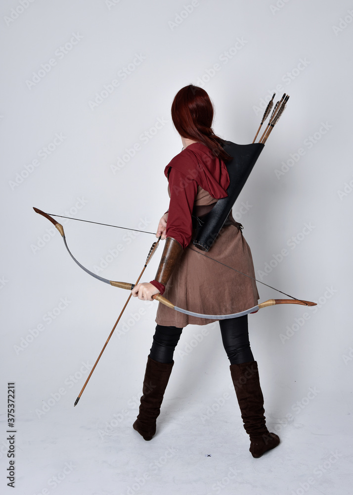 Wall mural Full length portrait of girl with red hair wearing  brown medieval archer costume.. Standing pose with back to the camera holding a bow and arrow,  isolated against a grey studio background.