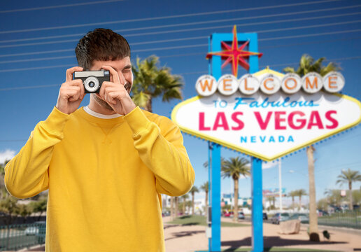 travel, tourism and photography concept - happy young male photographer in yellow sweatshirt taking picture with vintage film camera over welcome to fabulous las vegas sign background