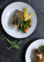 Grilled fish with lemon, herbs and rosemary. Fried fish fillets in a white plate. Beautiful presentation of food. Rucolla salad and carrots. Top view. Dark background