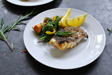Grilled fish with lemon, herbs and rosemary. Fried fish fillets in a white plate. Beautiful presentation of food. Rucolla salad and carrots.