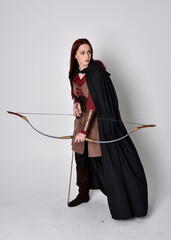 Full length portrait of girl with red hair wearing medieval archer costume with black cloak. Standing pose with back to the camera holding a bow and arrow,  isolated against a grey studio background.