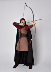 Full length portrait of girl with red hair wearing medieval archer costume with black cloak....