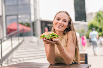 Young woman holding and eating eating tasty grilled burger outside in cafe