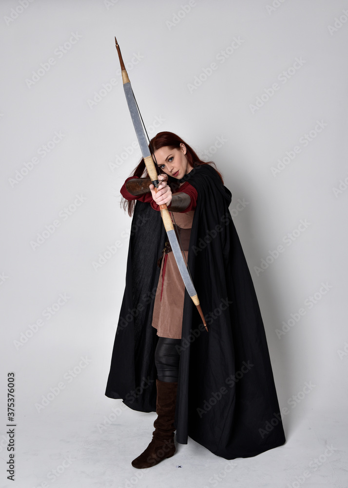 Poster full length portrait of girl with red hair wearing medieval archer costume with black cloak. standin