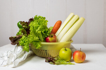 Vegetables in a plastic basket on a white table: carrots, celery, cucumbers, zucchini, apples, cabbage, lettuce. Healthy food concept