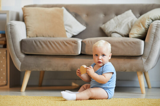 Horizontal Long Shot Of Playful Baby Sitting On Floor In Living Room Playing With Toy Looking At Camera