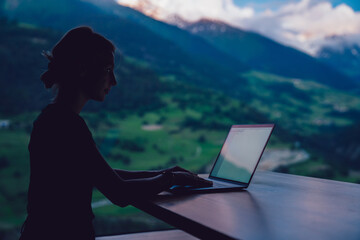 Skilled female digital nomad working remotely on freelance project sitting at table near big window...