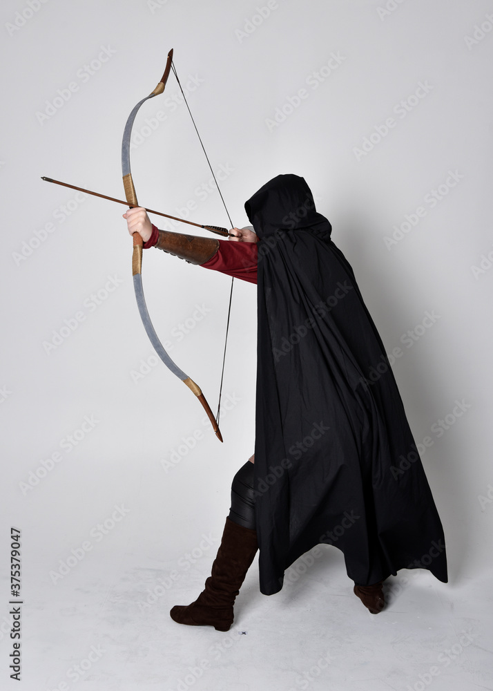 Poster Full length portrait of girl with red hair wearing medieval archer costume with black cloak. Standing pose with back to the camera holding a bow and arrow,  isolated against a grey studio background.