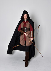 Full length portrait of girl with red hair wearing medieval archer costume with black cloak. Standing pose with back to the camera holding a bow and arrow,  isolated against a grey studio background.