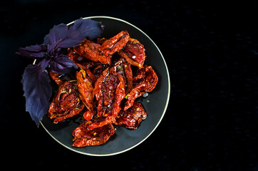 top view of plate with sun-dried tomatoes in olive oil, with spices and basil leaves on dark background with copy space, place for text