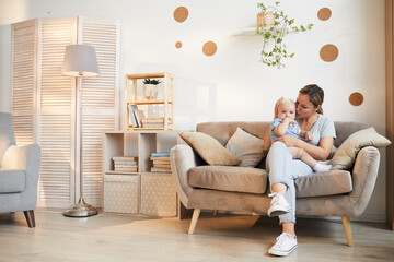 Horizontal long shot Caucasian woman wearing casual outfit sitting on sofa with her kid on lap, copy space