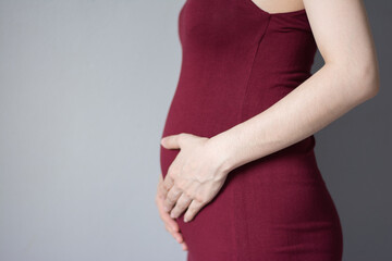 Pregnant woman in dress holds hands on belly on a gray background.