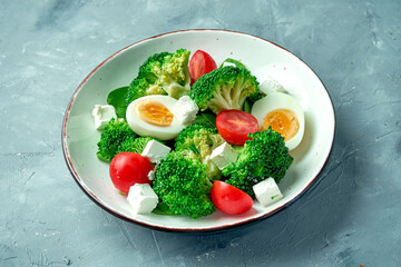 Fresh and healthy salad with broccoli, egg, feta cheese and tomatoes in a white bowl on a gray background