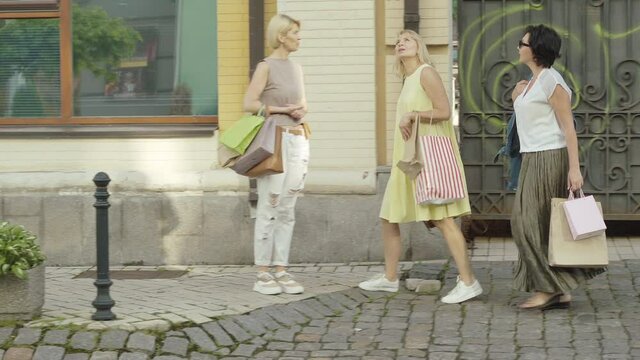 Wide Shot Side View Of Three Shopaholics Searching For Shop On Sunny Summer Day. Portrait Of Carefree Mid-adult Rich Ladies Walking Along City Street With Shopping Bags And Talking.