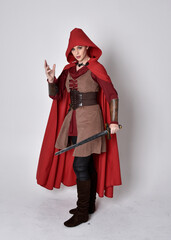 Full length portrait of girl wearing medieval costume and red cloak. Standing pose holding a sword,  isolated against a grey studio background.