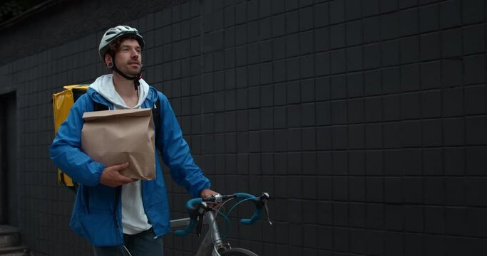 Male courier in protective helmet with food transportation bag carrying paper package and pushing bike at street. Man smiling and walking while delivering order to customer