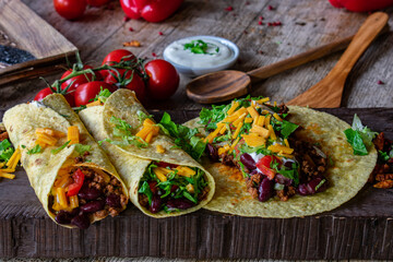 Burritos with ground beef filling, kidney beans, cheddar cheese, tomatoes and lettuce on wooden board