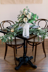 A beautiful square wood table with two chairs at a wedding reception decorated with a pretty bridal bouquet with white roses and lots of eucalyptus leaves and greenery. 
