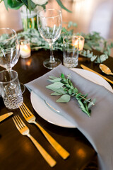 A beautiful wedding reception table setting with eucalyptus leaves on top of gray napkins, wine glasses and votive candles are in the background with gold silverware - fork, spoon, and dinner knives. 