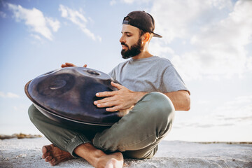 close up photo of stylish bearded man holding a unique handmade musical instrument handpan playing...
