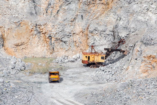 Stone Open Pit Mine. Excavator Is Loading Raw Materials Into Yellow Truck For Transport To Combine