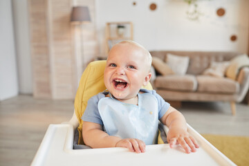 Horizontal portrait of cheerful little child sitting on high chair with food around his mouth smiling at camera, copy space