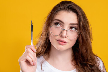 Curious woman portrait. Motivation inspiration. Smart pensive lady in glasses pointing up with pen isolated on yellow copy space background. Knowledge education. Study learning.
