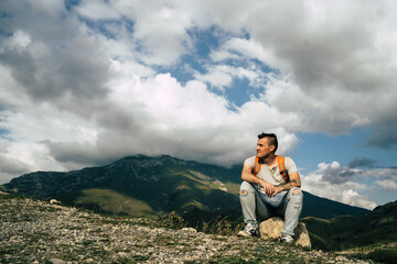 Naklejka premium Young man sitting on hill. Dreamy male sitting on altitude and looking at Peaks of magnificent rocks located against bright cloudy sky