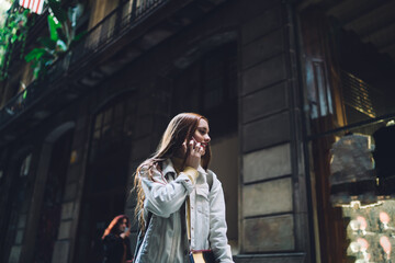 Joyful modern female student talking on smartphone while walking along street