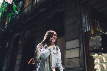 Fototapeta premium Joyful modern female student talking on smartphone while walking along street