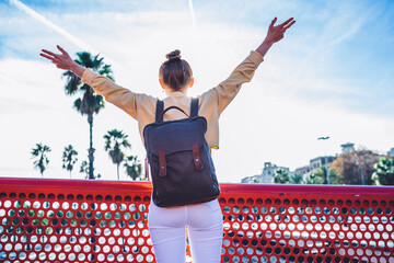 Fototapeta na wymiar Back view of female traveler with trendy touristic backpack raising hands in air while feeling freedom during getaway vacations for inspiration and recreation, millennial wanderer enjoying own life