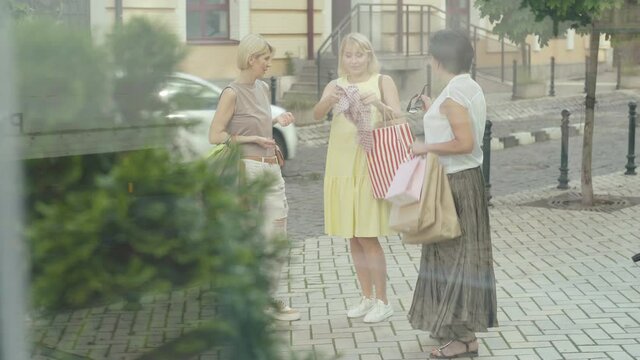 Elegant Lady Bragging New Stylish Summer Scarf To Friends On Urban Street. Wide Shot Portrait Of Three Happy Caucasian Mid-adult Women Enjoying Shopping In City. Shooting Behind Glass.