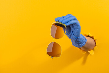 Hand in a latex glove giving sunglasses through torn yellow wall