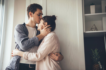 Cute young couple kissing in the kitchen and embracing near the window
