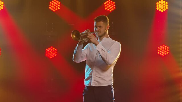 A young man in a white shirt and trousers plays the trumpet. Picture taken in a dark studio with red spotlights and smoke in the background. Slow motion. Close up.