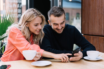 Cheerful caucasian woman and man couple in love browsing web page on smartphone making booking, 20s smiling female watching video on mobile phone of her boyfriend resting in cafe on break together