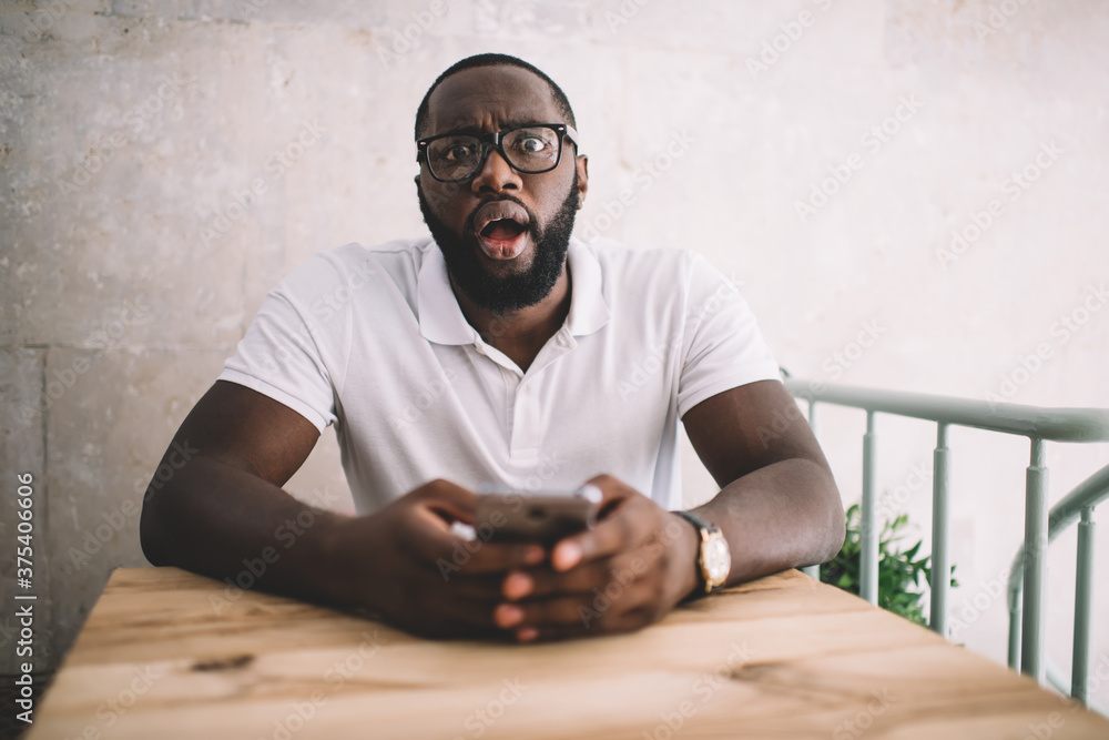 Poster portrait of amazed black blogger in optical spectacles for eyes correction holding modern cellphone 