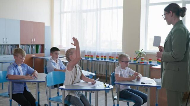 teacher shows cards with assignment for pupils, children raise hands knowing the correct answer while sitting at their desks at school
