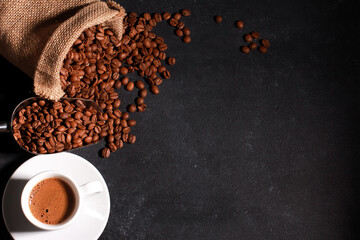 White cup and brown coffee beans in metal scoop scattered from burlap bag on dark table background with copy space. Arabica grains, top view. Coffee shop, caffeine, roast concept