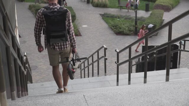 Man walking with folded electric scooter up stairs in city near modern building. Ecological technological lifestyle. E-Mobility. Person Carrying Electric Scooter Folded Position. Eco urban transport