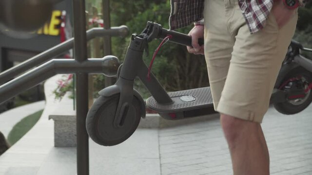 Young caucasian man in casual clothes with a backpack carries a folded electric scooter in his hand up the stairs of an office building. Man Carrying E-Scooter in folded position From Work