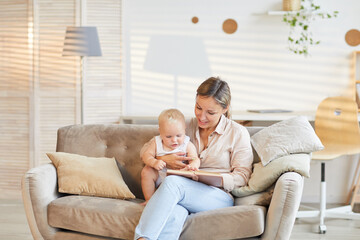 Modern young mother sitting on sofa in living room with her curious kid reading book, copy space