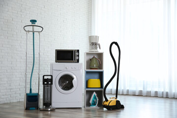 Modern vacuum cleaner and different household appliances near window indoors