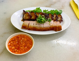 Crispy Roasted Crackling Pork Belly in Chinese Cantonese cuisine called Siu Yuk, fresh herbs, chili garlic dipping sauce, white plate, marble table top. 