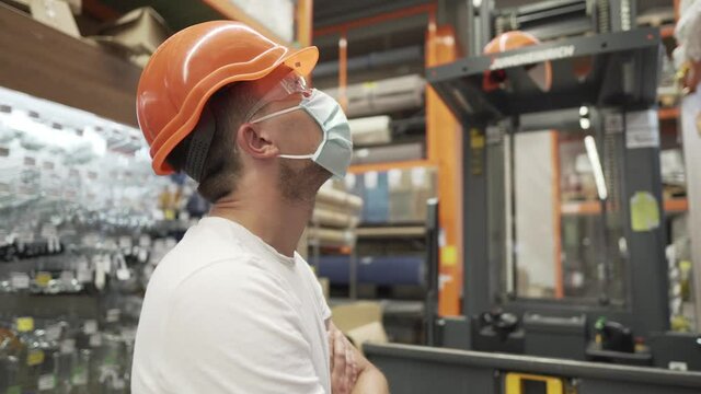 Man working in a warehouse, inventory of a store, and operates a forklift. A construction supermarket worker wearing an orange helmet, goggles and a mask during the coronoviris and covid 19 pandemic